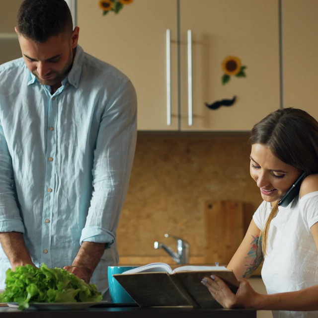 Pareja en la cocina, uno lee y el otro prepara la comida